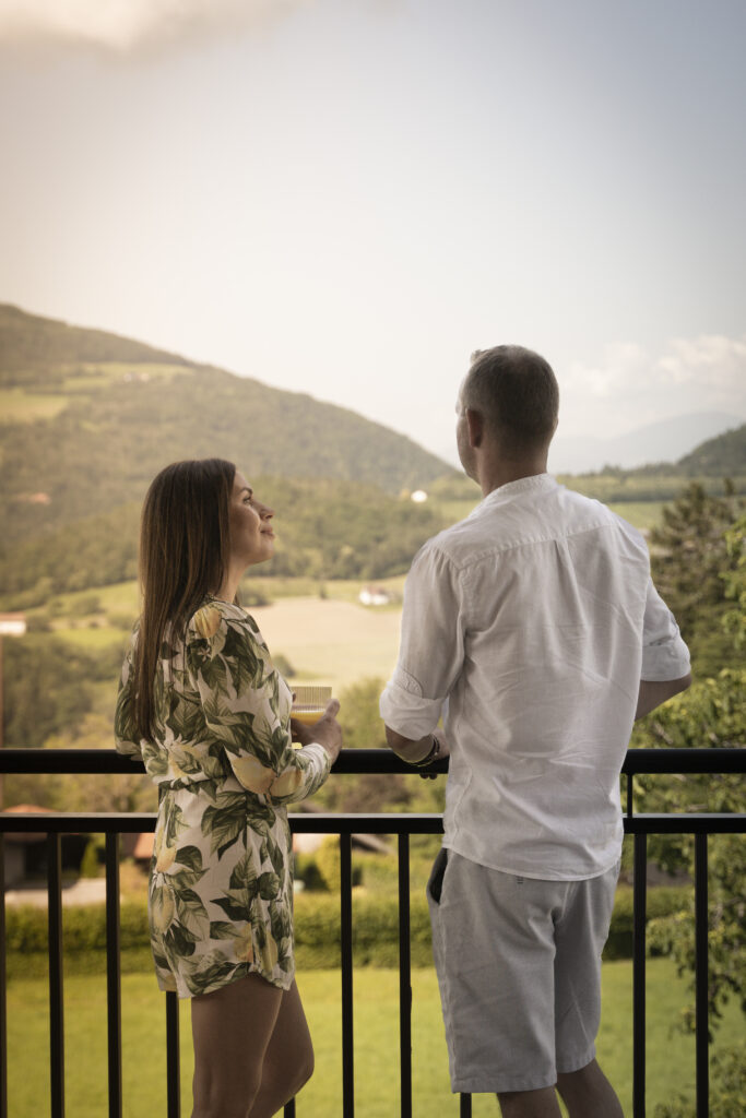 Zwei Gäste am Balkongeländer, davor die Hügel, Wiesen und Felder der Oststeiermark
