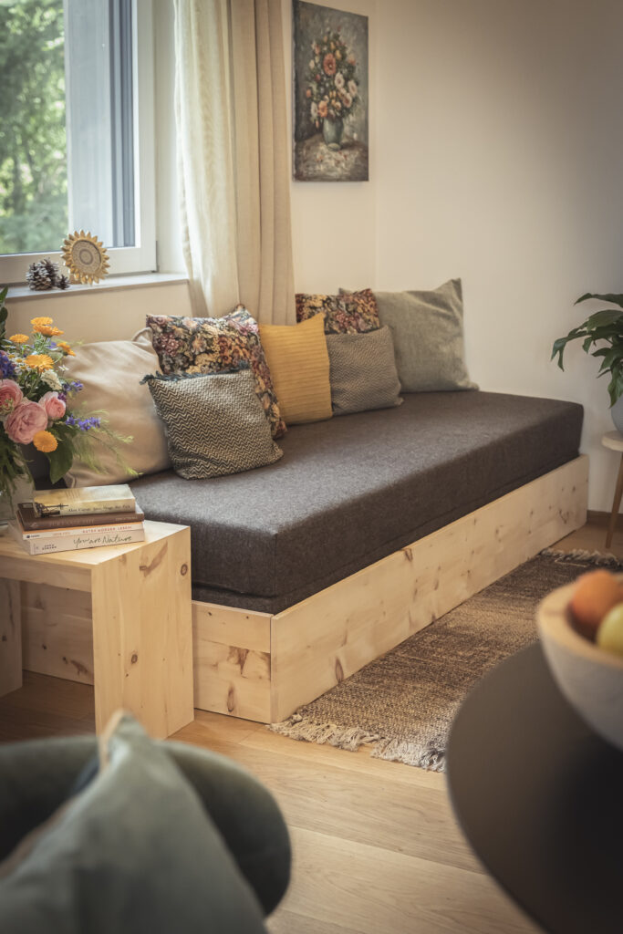 Sitzbank aus Massivholz mit Kissen vor dem Fenster, daneben Blumen und Bücher auf einem Hocker